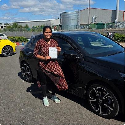 Woman standing by a black car in a car park, holding a document and smiling.